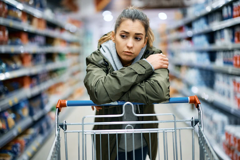 Young woman leaning on a grocery cart in a supermarket aisle looking worried