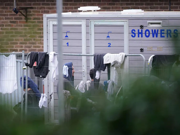 Migrants queue outdoors for a temporary shower facility, behind a wire fence where clothes are hanging