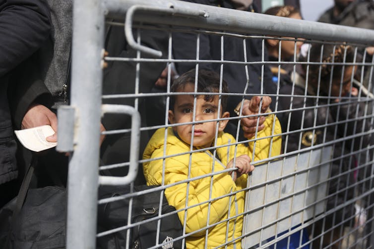 A young boy in a yellow jacket behind a fence. He is holding an adult's hand.