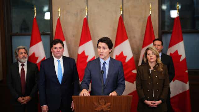 A man with dark hair surrounded by three men and a woman stands in front of a podium speaking. A row of Canadian flags are behind him.