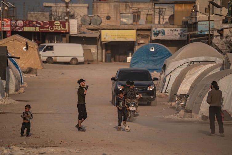 Children play soccer on a dusty street with tents.