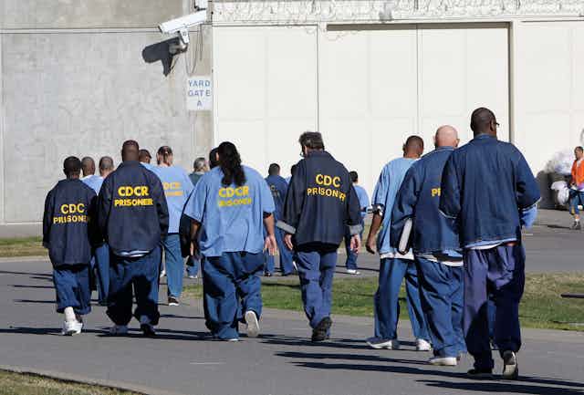 A group of mostly Black men walk away from the camera in blue prison uniforms.