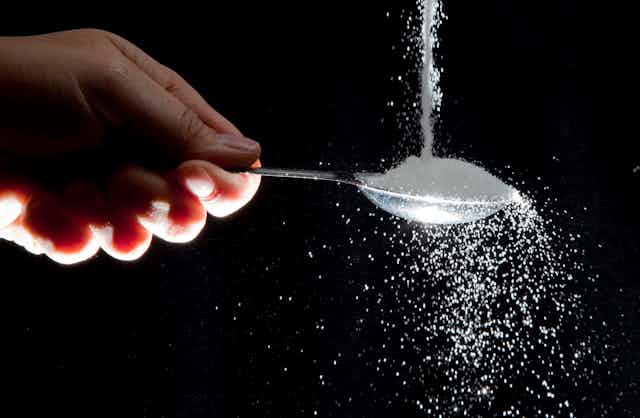 A hand holds a spoon as sugar pours onto it