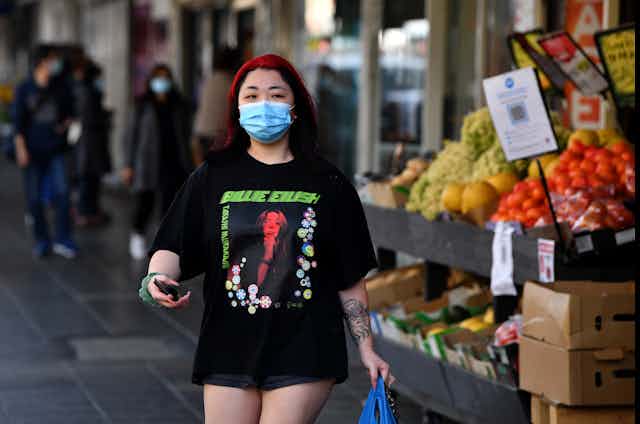 Young woman wearing mask holding phone and shopping bag outside grocery shop