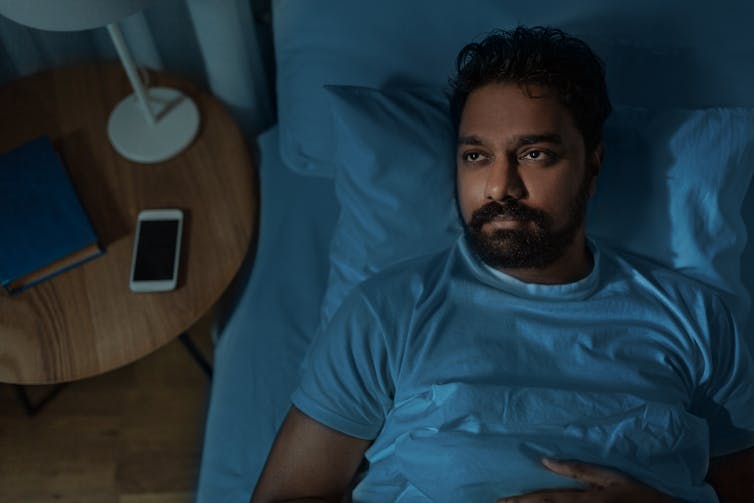 man lies awake in bed at night with mobile phone next to him on bedside table