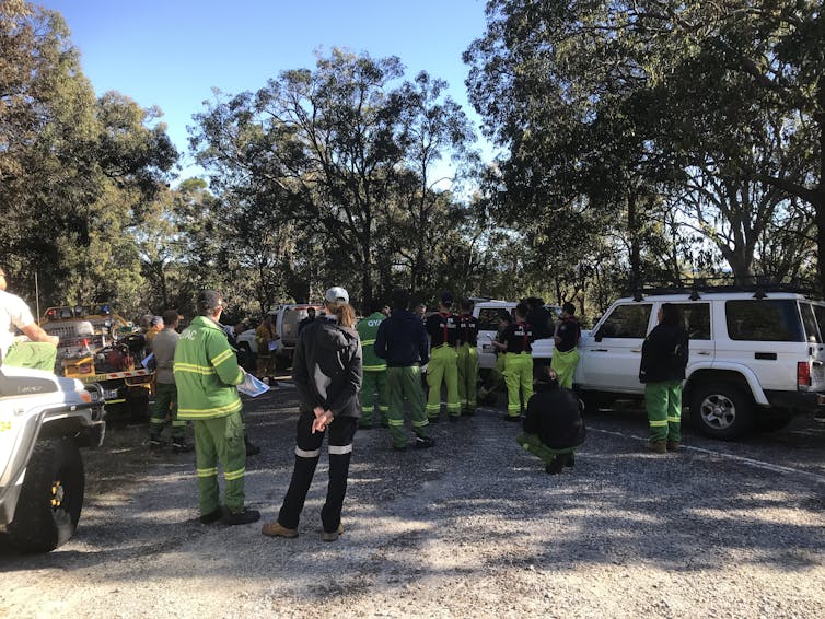 Large group of people from various agencies, in uniform, standing at an intersection for a pre-burn meeting. With vehicles nearby