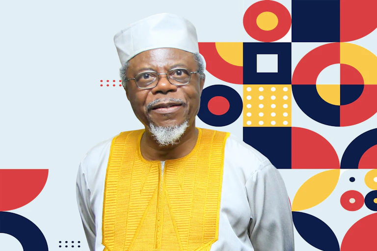 A man with spectacles and a grey beard and moustache poses for a photo in traditional Nigerian attire against a backdrop of bright African patterns in orange, yellow and black.