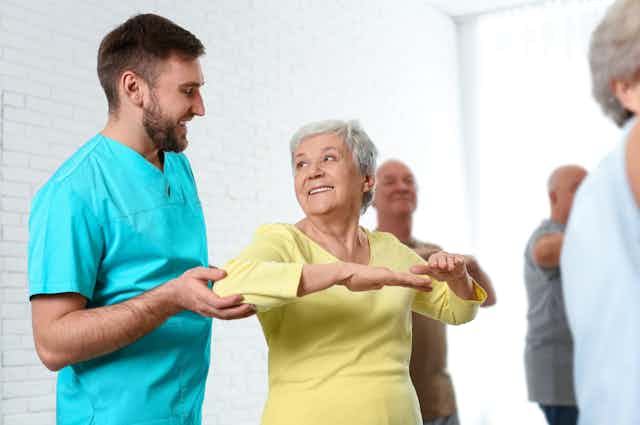 Mujer mayor haciendo gimnasia mientras un trabajador asistencial le da indicaciones.