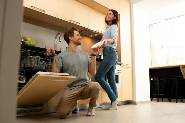 Smiling male is putting dishes into dishwasher, chatting with woman in the background, at home, kitchen.
