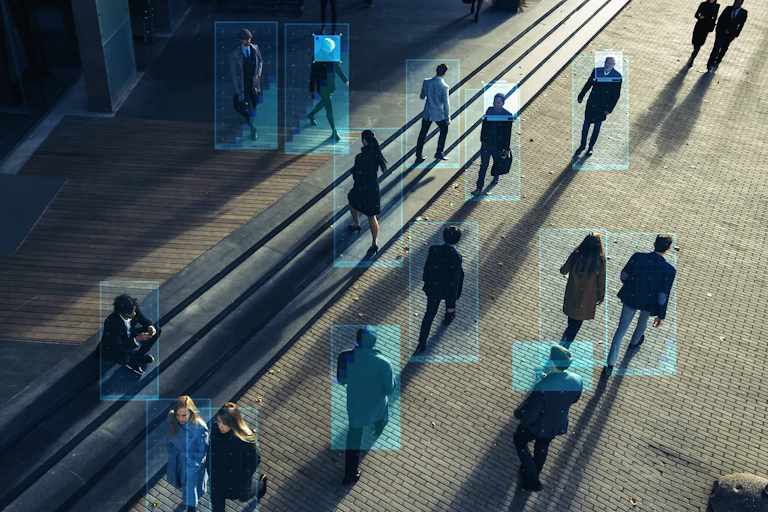 A number of human beings walk in different directions. The angle shows them from above and they are casting long shadows. Blue digital boxes frame each figure.