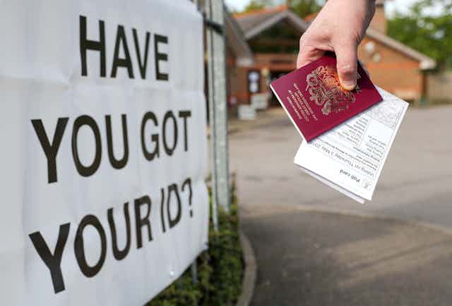 A hand holding a polling card and a passport in front of a sign rhave you got your ID?'eading '