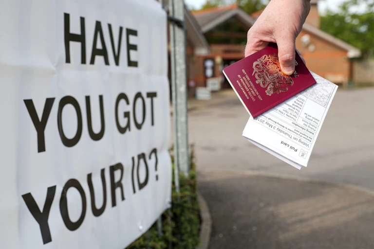A hand holding a polling card and a passport in front of a sign rhave you got your ID?'eading '