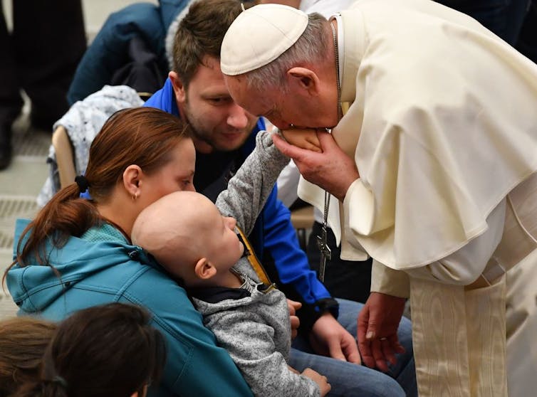 The pope, wearing a white skullcap, bends down to kiss the hand of a child in his mother's lap.