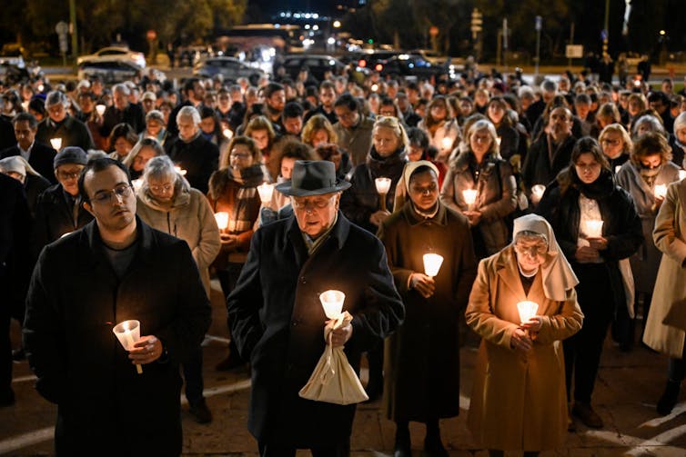 A crowd of people in winter clothing holding candles during a nighttime vigil.