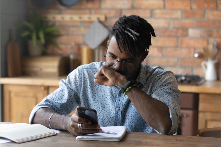 A young Black man sits at a desk using a smartphone.