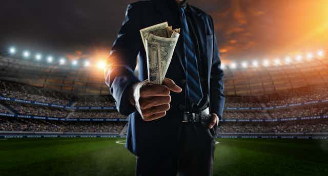 A man wearing a blue sport coat and tie holds a wad of cash while standing on the field of a baseball stadium at twilight.