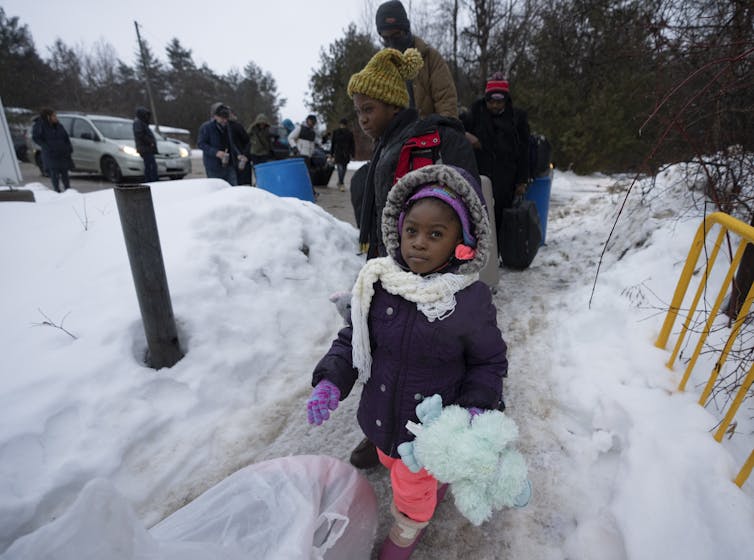 A young Black girl in a snowsuit carrying her belongings stares at the camera.