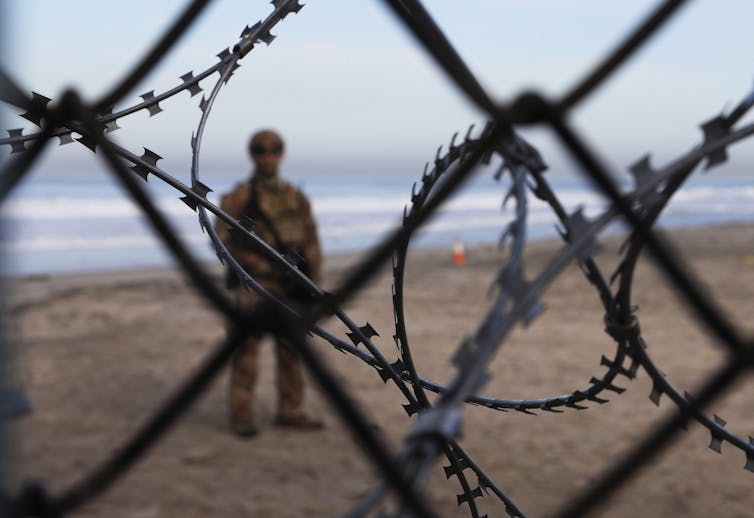 A heavily armed Border Patrol agent is seen through a chain link fence strewn with barbed wire.