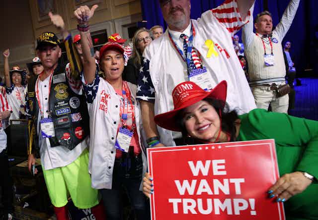 People pose for a photo wearing colorful pins and one woman holds a sign that says 'We want Trump!'