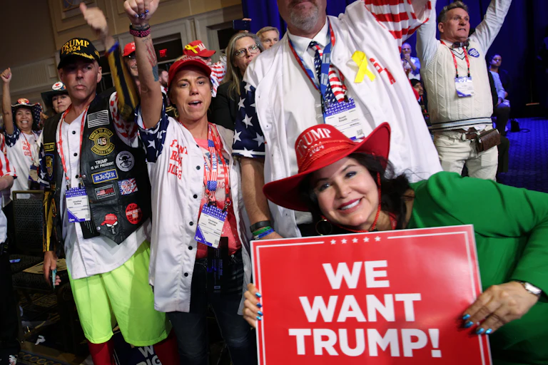 People pose for a photo wearing colorful pins and one woman holds a sign that says 'We want Trump!'