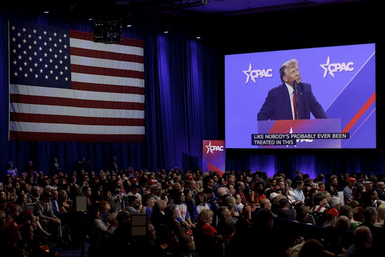 A large crowd of people look toward a screen that shows a white man in a dark suit. Next to the screen is a large American flag