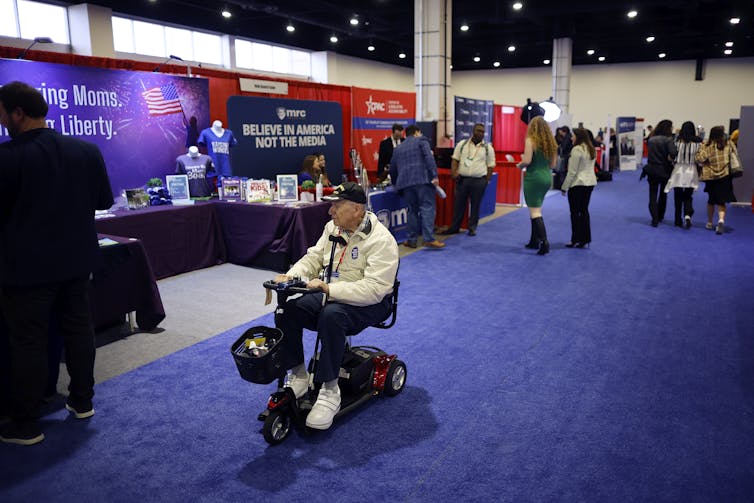 A man in a wheelchair goes past a booth in a convention room that says 'Believe in America, not the media.'