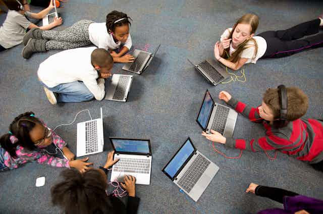 a group of kids lie in a circle on the floor using laptop computers