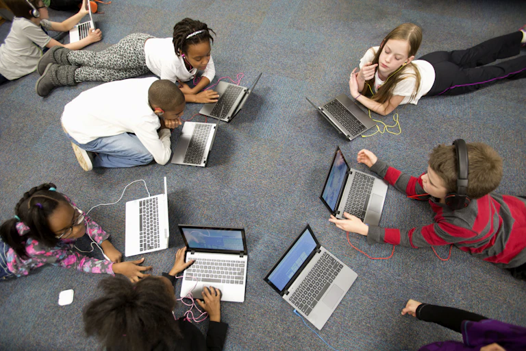 a group of kids lie in a circle on the floor using laptop computers