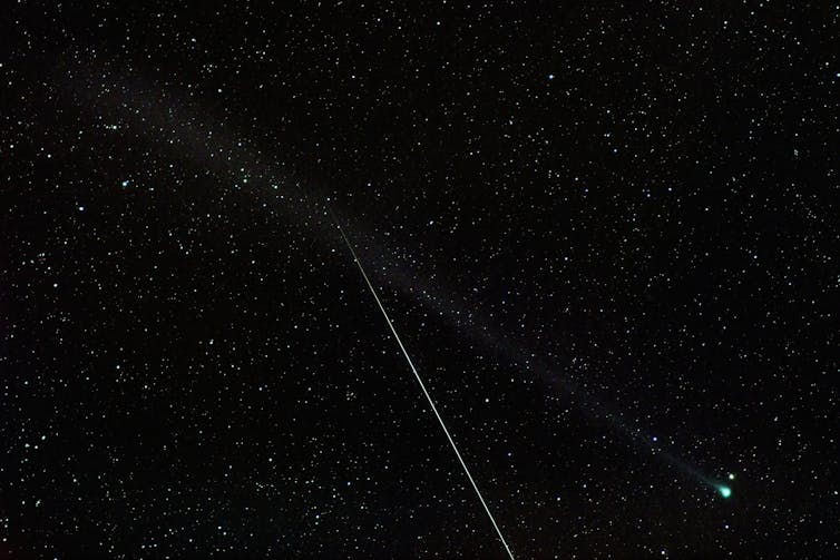 A comet sits with its greenish head at the bottom right of the image, with its tail extending to top left. In the foreground, a bright Eta Aquariid meteor can be seen, descending nearly vertically from the middle of the frame