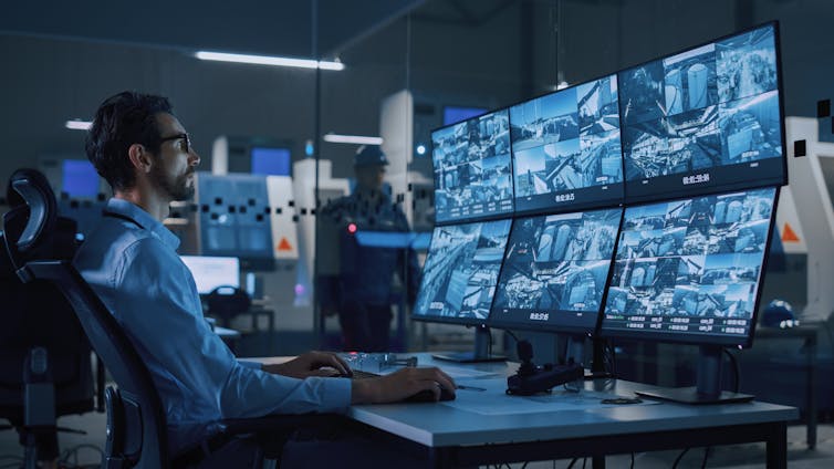 A man sits at a desk in front of a bank of screens, each showing footage from CCTV cameras.