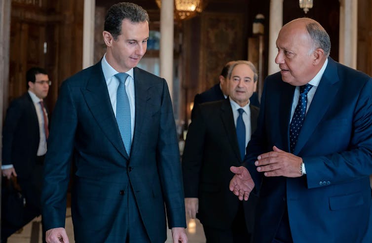 Syrian president Bashar al-Assad with the Egyptian foreign minister, Sameh Shoukry wearing suits and ties. A aide watches from behind.