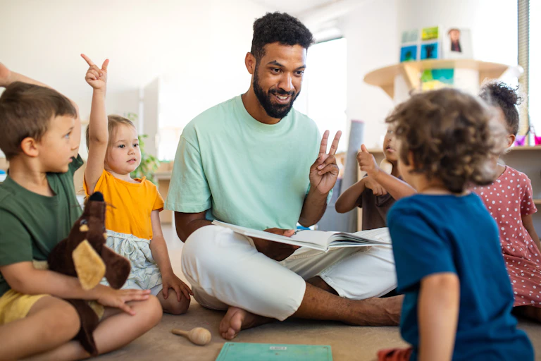 Group of small children with male teacher or childcare worker, sitting on floor indoors, learning.