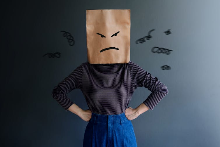 Woman with her hands on her hips and a paper bag over her head with an angry face drawn on it