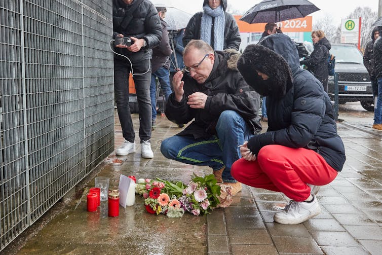A man and woman in winter clothes crouch by a makeshift memorial with candles and flowers.