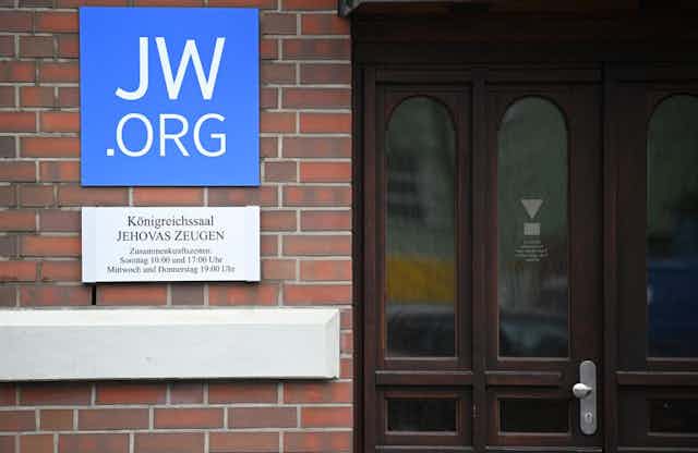A blue sign that says 'JW.ORG' in large font on a brick wall next to a wooden door.