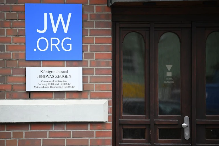 A blue sign that says 'JW.ORG' in large font on a brick wall next to a wooden door.