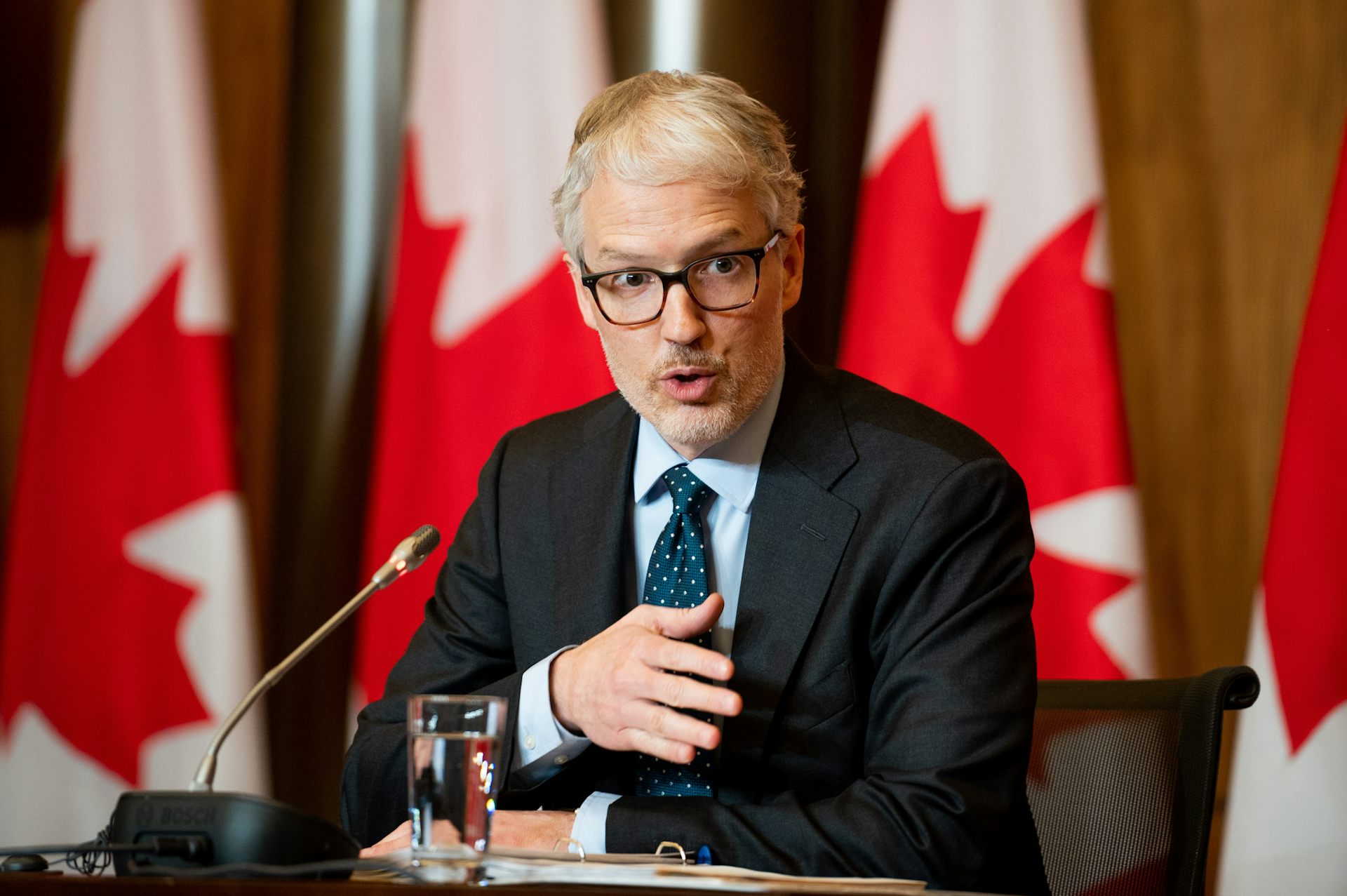 A man wearing a suit and glasses speaks into a microphone from behind a desk. Canadian flags stand in the background.