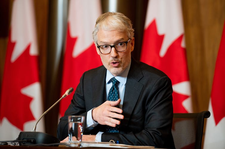 A man wearing a suit and glasses speaks into a microphone from behind a desk. Canadian flags stand in the background.