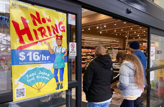 a group of people walk into a store with a yellow window poster stating 'Now Hiring'.