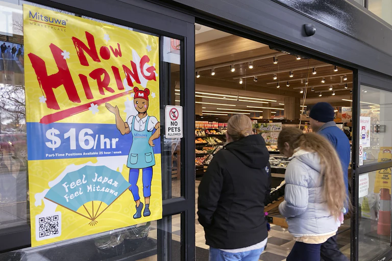 a group of people walk into a store with a yellow window poster stating 'Now Hiring'.