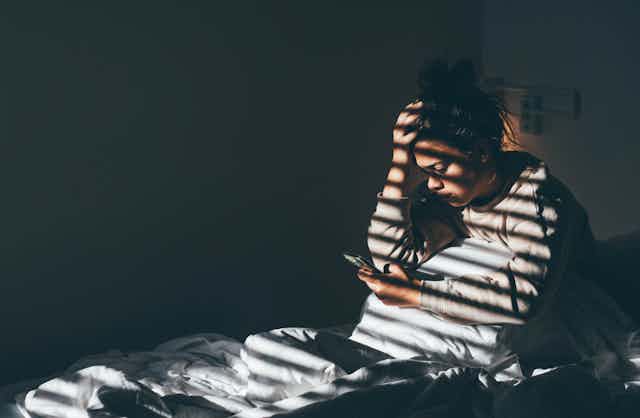 Young woman sitting on bed in the darkness and staring at her phone, with light shining through window.