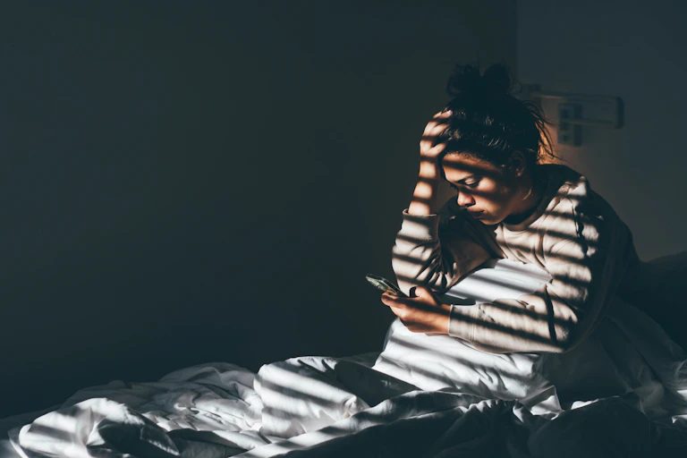 Young woman sitting on bed in the darkness and staring at her phone, with light shining through window.