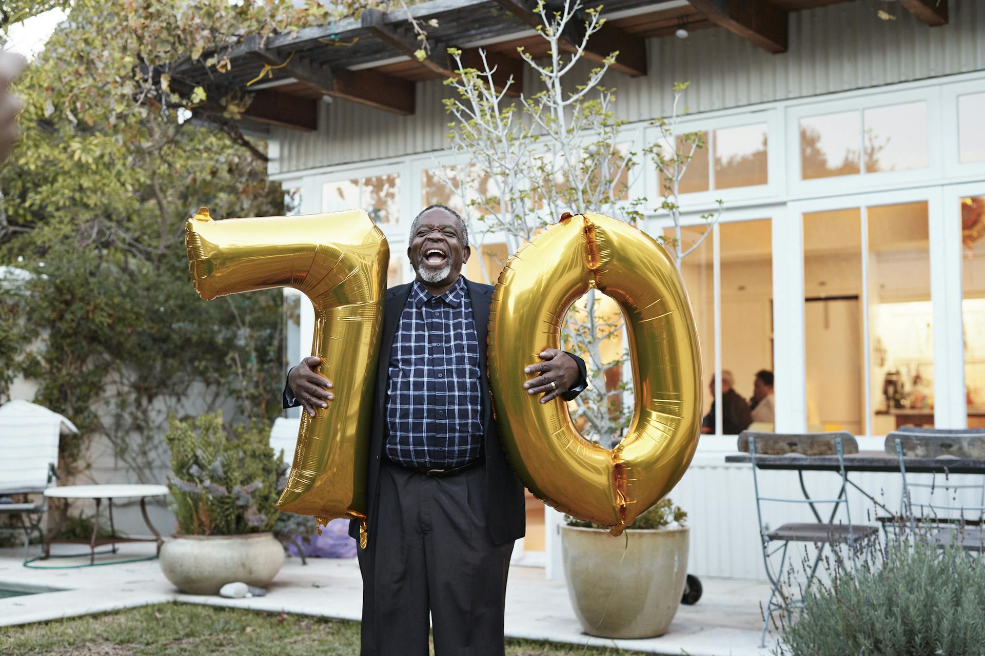 Older adult holding gold balloons of the number 70 in a backyard