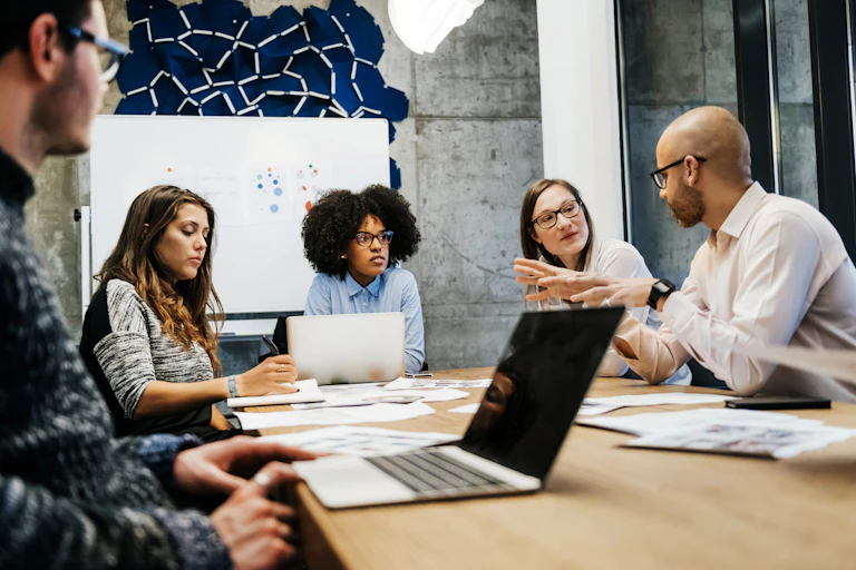 Three women and two men sit at a table with laptops and papers on it. A whiteboard is in the background. They are talking.