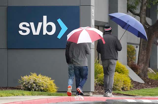 Several people carrying umbrellas walk in rain past wall with sign that reads svb