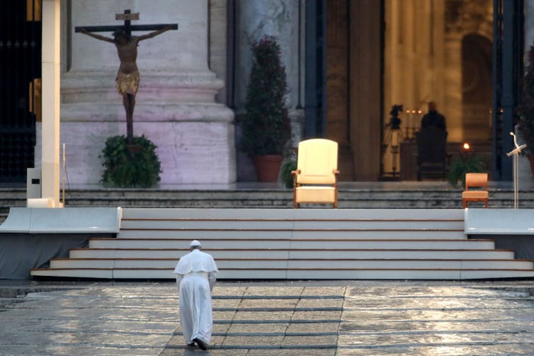 Man in a white robe walking towards a set of stairs, a single white and gold seat is at the top of the stairs and a crucifix hangs on the wall behind it.