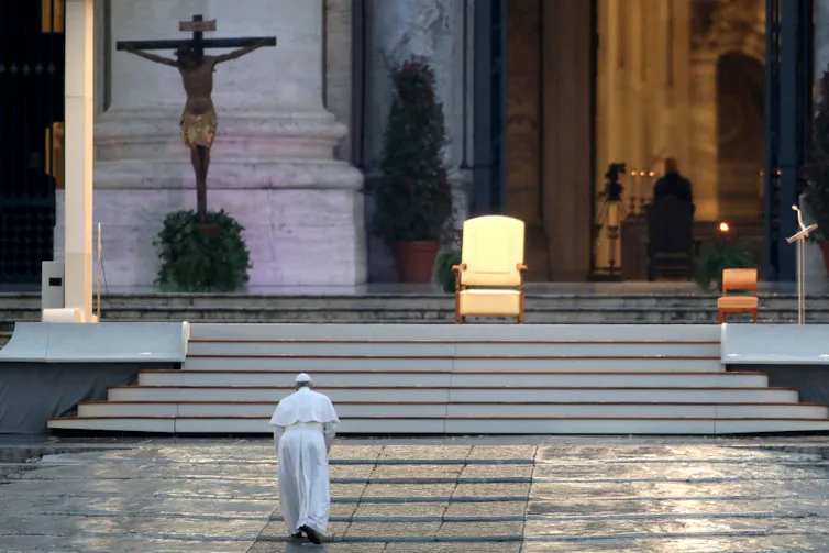 Man in a white robe walking towards a set of stairs, a single white and gold seat is at the top of the stairs and a crucifix hangs on the wall behind it.