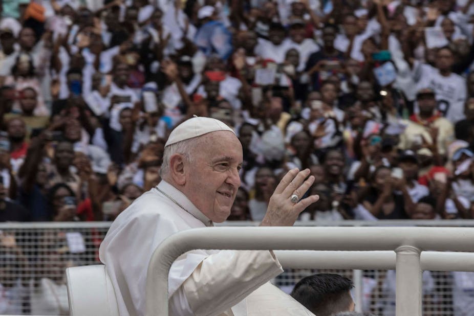 Man in a white robe and white cap sitting in the back of a car lifts his hand in greeting - a crowd of people in the background waves back