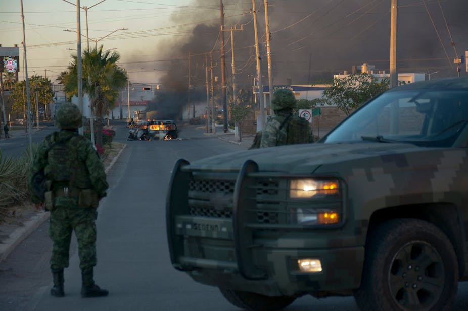 Dos soldados vestidos de camuflaje, de pie frente a un vehículo todoterreno sucio, miran un vehículo en llamas en medio de una calle, con humo que se eleva en el aire. 