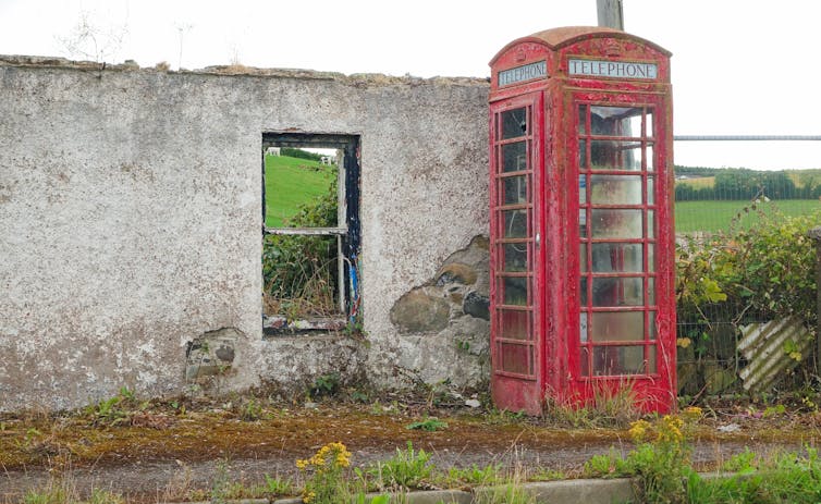 A derelict red telephone booth next to the sole remaining wall of a building in a rural area